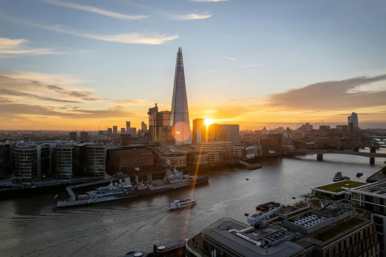 the sun is rising over the city as buildings and boats stand on the river bank