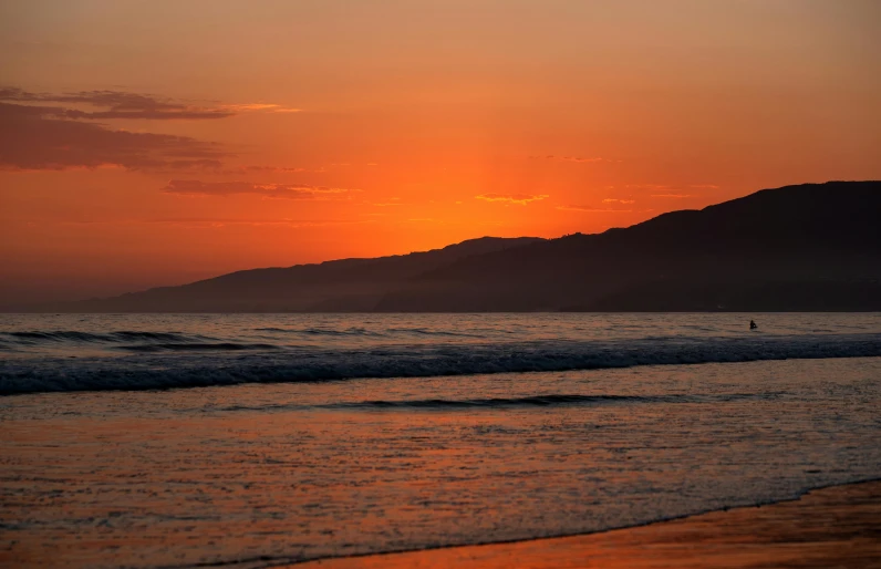 a person on a surfboard in the distance is on a calm beach at sunset