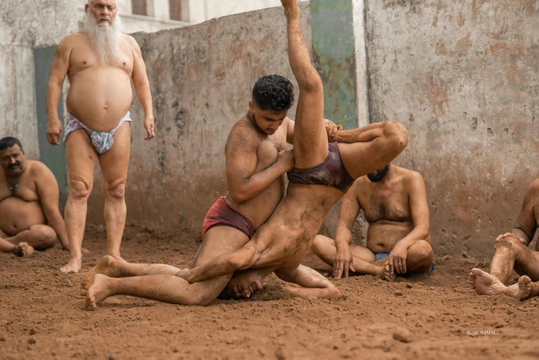 four men in swim trunks and , in front of an old stone building one standing and another squatting