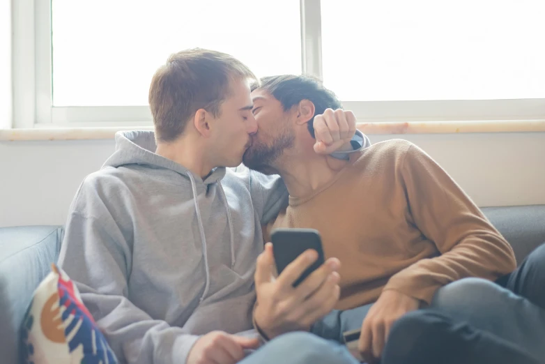 a couple sitting on a couch one kissing another as he holds a cell phone