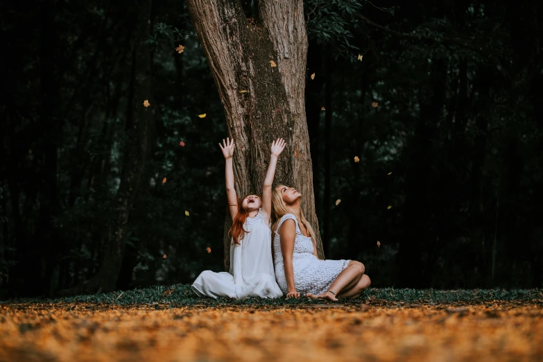 two women sitting under a tree with their hands up in the air