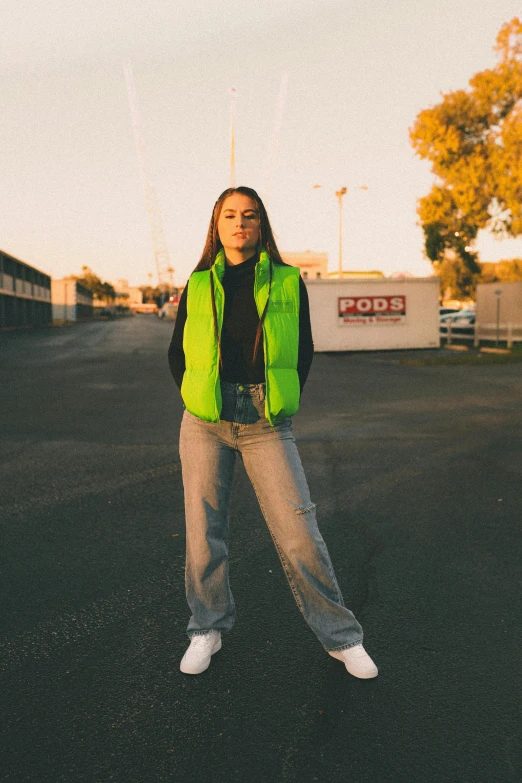 a woman stands in the middle of an asphalt area while wearing a neon vest and white sneakers