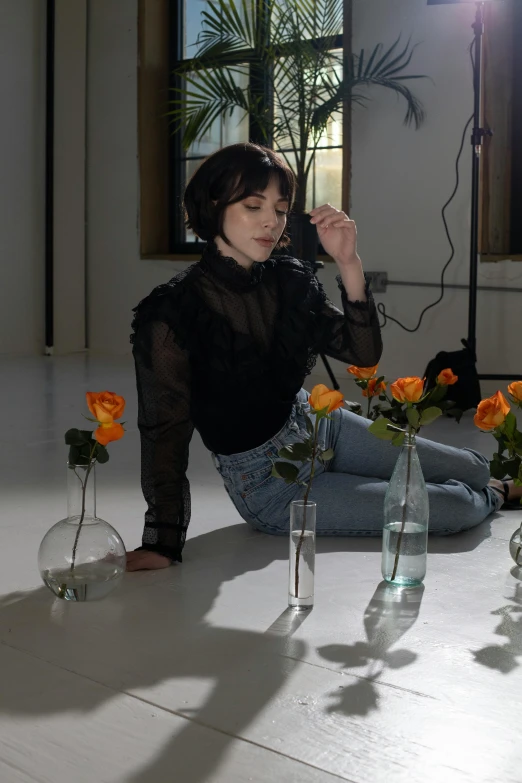 a woman in jeans and a sheer shirt crouches near a table of orange roses