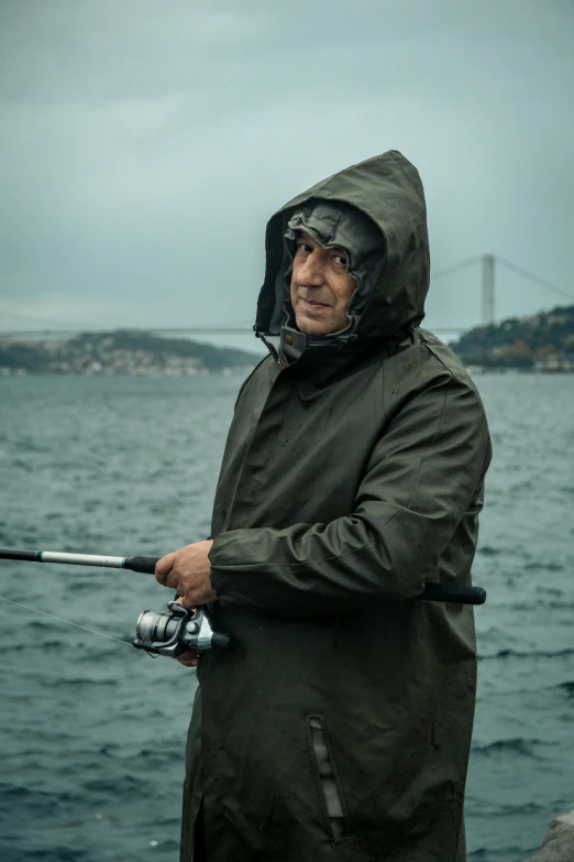 an older man with a hood on, holding a fishing rod by the water