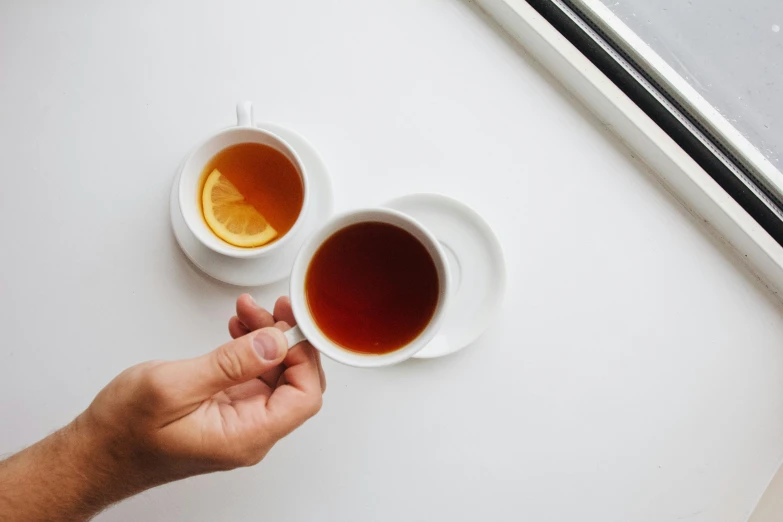 two cups of tea sit on a desk near windows
