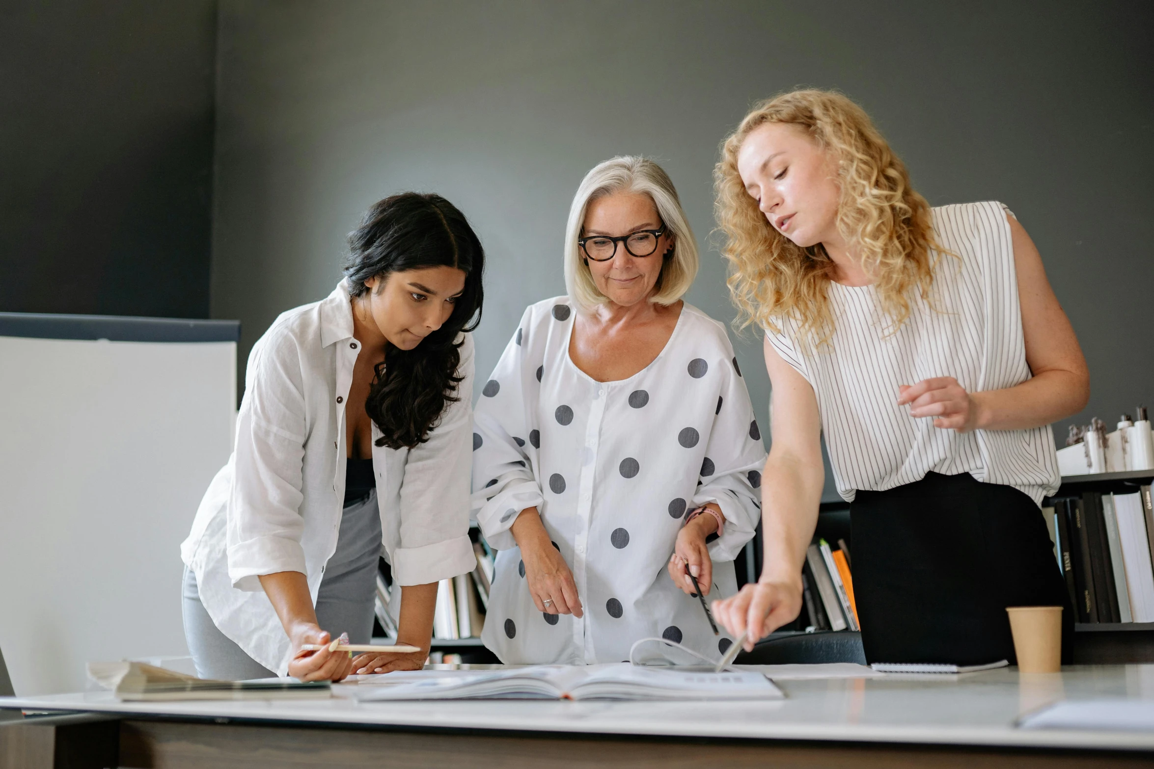 three women standing around a desk looking at a book