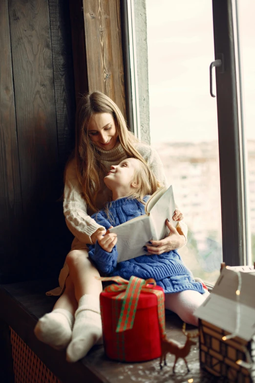 mother reading book to daughter on window sill