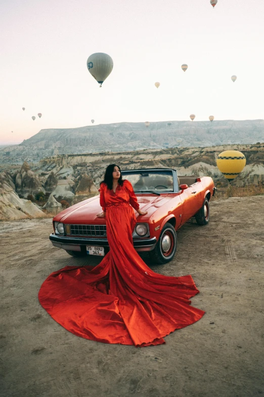 woman in red dress standing by classic car with balloons flying above