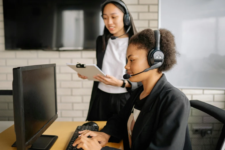 the young woman has her hands under her chin while talking on the phone and standing behind a computer with the other person in front