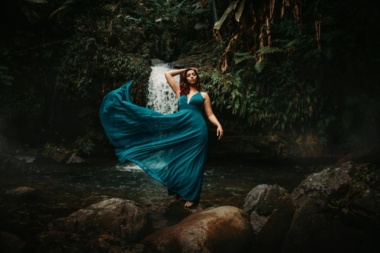 a woman wearing a blue dress standing on rocks in front of a waterfall