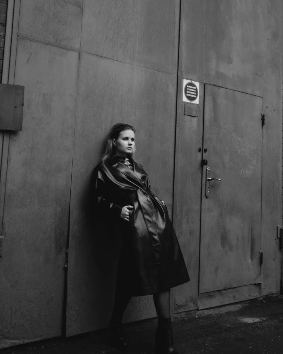 a woman wearing an oversize coat and heels is leaning against the wall