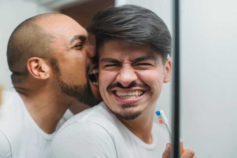 two males in white shirts are smiling with one brushing his teeth