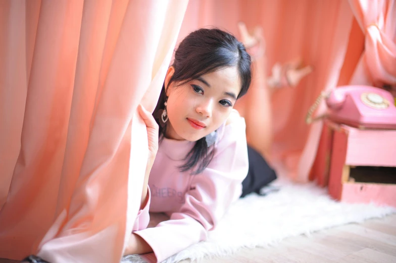 an asian woman sitting down leaning against the curtain of her home