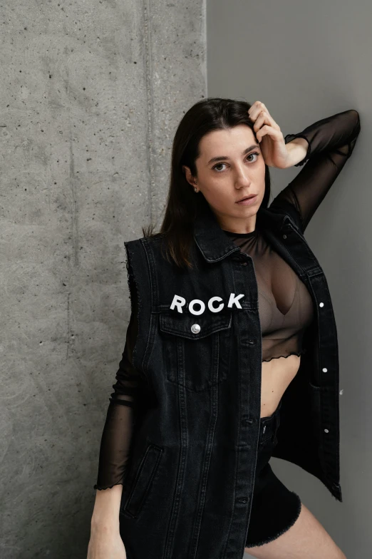 a beautiful young woman leaning on a wall wearing a sheer shirt and jean vest