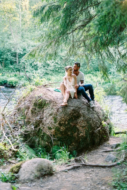 two people sit together on a rock near a river