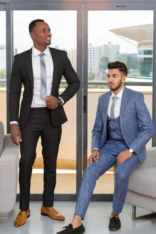 two men stand in front of glass doors, and one man in a blue suit has his hand on his chin