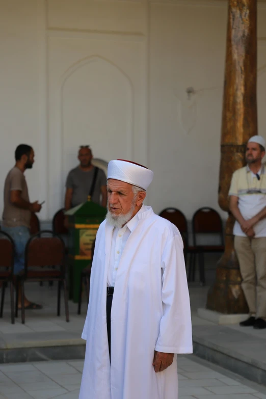 older man wearing white linen dress standing in front of two seated men