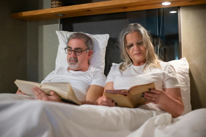 a man and woman sit on a bed with white sheets reading