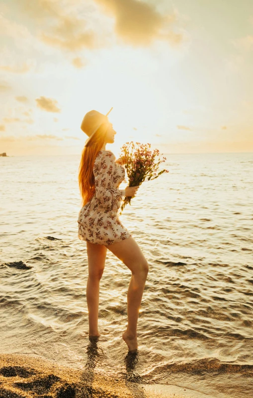 woman in floral dress holding up plants by the ocean