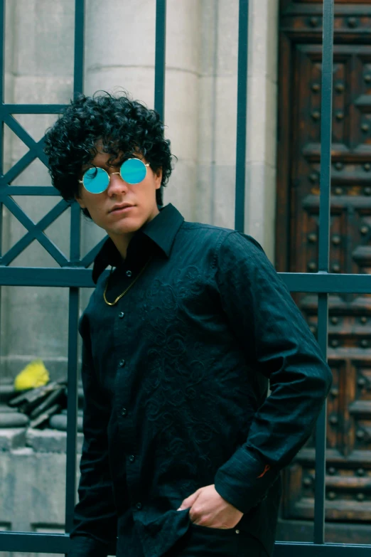 a person with curly hair and glasses standing by a gate