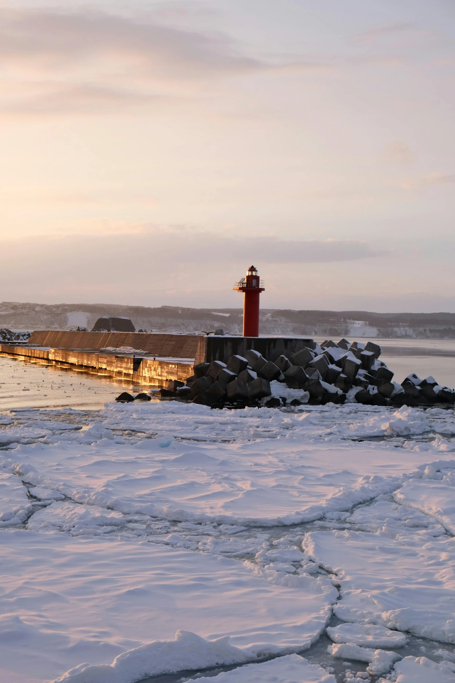 a small lighthouse is at the end of a long jetty that's off in the distance, with ice in the foreground