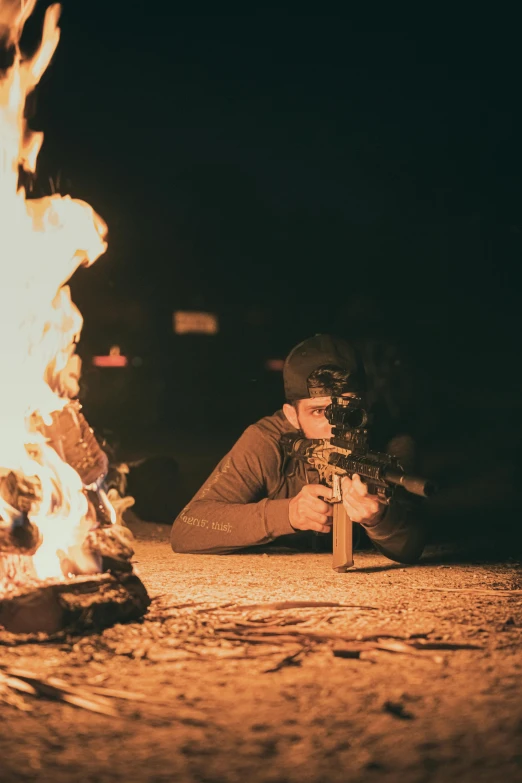 person laying down with a rifle near a fire