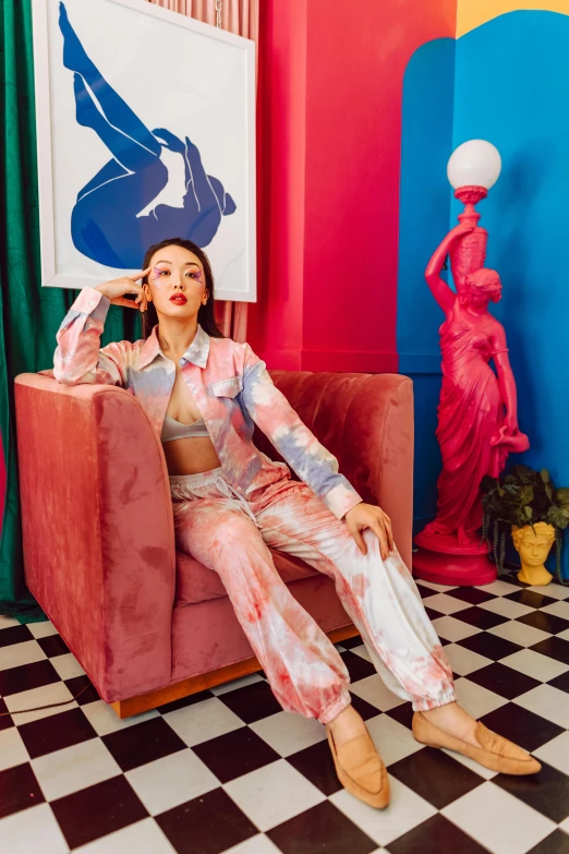 a young woman wearing a pink suit and tie dye pants is sitting on a red chair in front of a statue and artwork