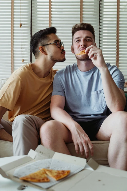 two guys sitting on a couch eating pizza together