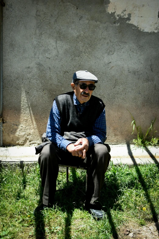 an older man with a vest and hat sits in the grass