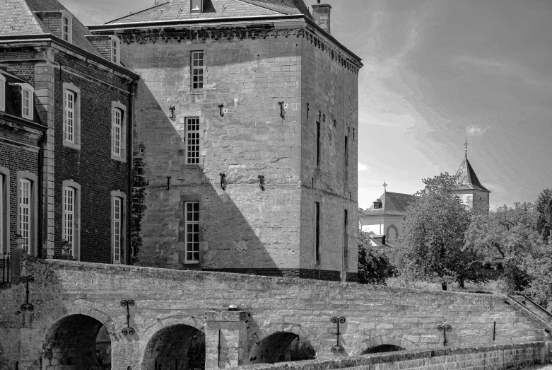 a black and white po of an old building in the foreground, with a large gate on the side, to the right and another one of it is a brick building with a clock tower on top and a bridge