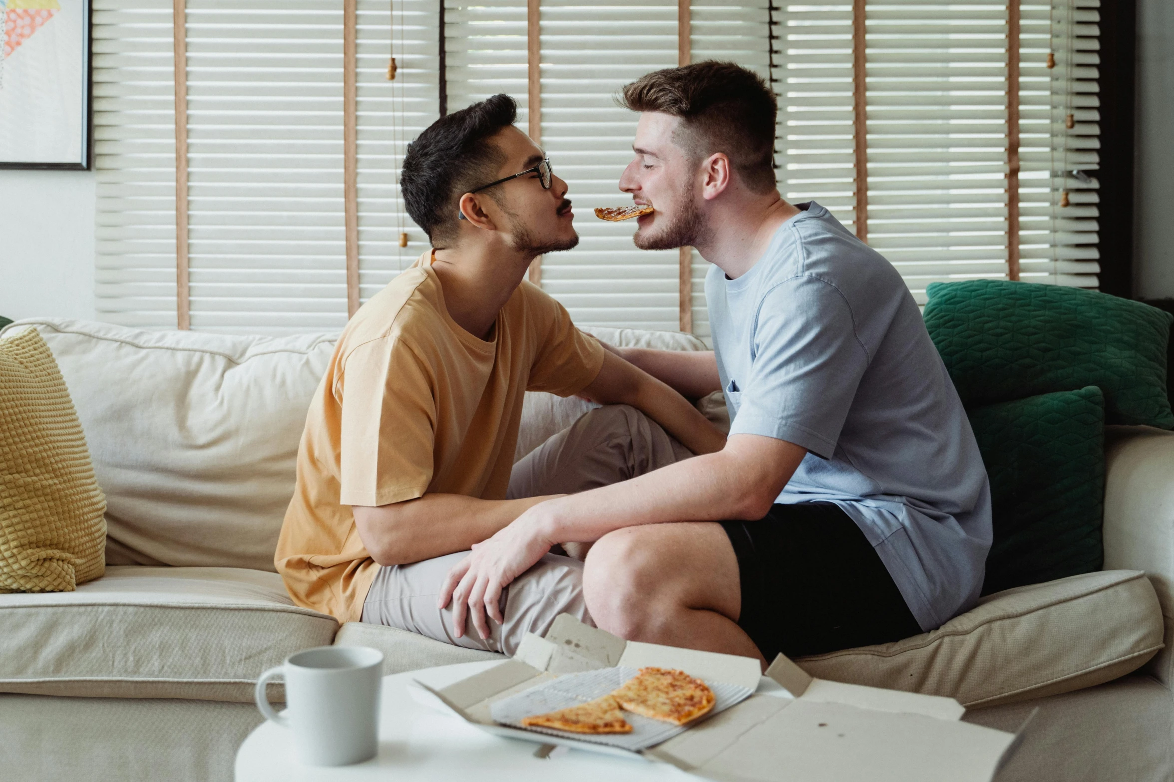 two young men sitting on top of a couch sharing pizza