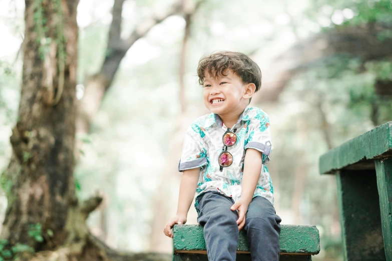 a child in a blue and white shirt sits on a green bench with trees behind him