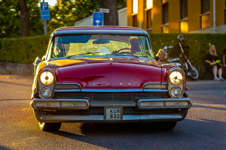 an old red car driving down the street with lights on