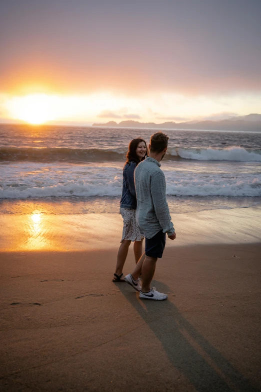 a man and woman standing on a beach with the sun setting