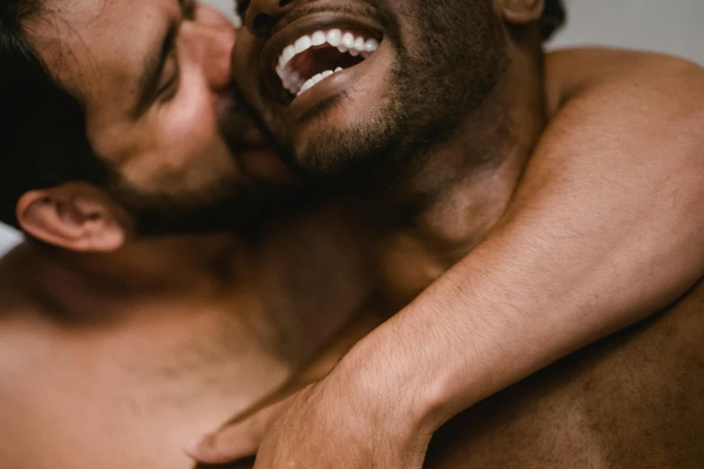 a shirtless man gets kissed by another man's cheek