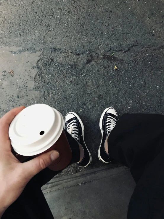 a persons hand holding a white coffee cup and black sneakers