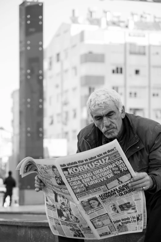 black and white image of a man sitting on a city ledge reading a newspaper