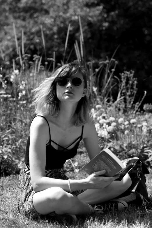 a woman wearing sunglasses sitting in grass reading