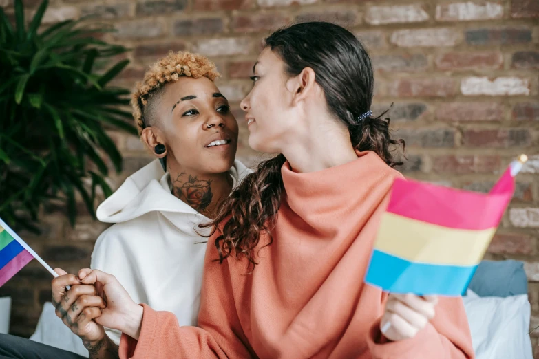 two women kissing holding rainbow colored flags in front of a brick wall