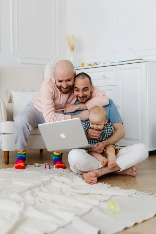 two men, a baby and a woman are sitting on a bed, both holding an apple laptop