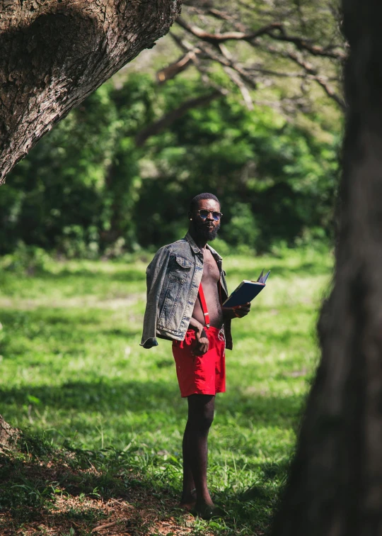 man in a red shorts wearing a grey and blue jacket with his arms in his pockets standing under a tree