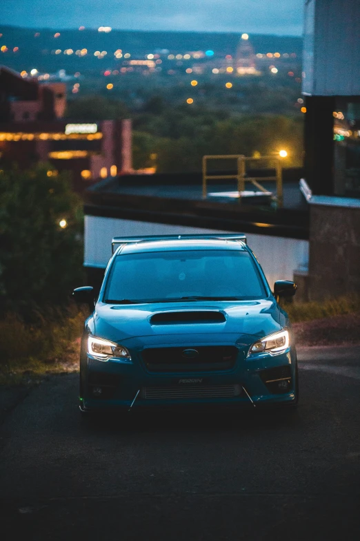 the front end of a blue sedan with city lights in the background