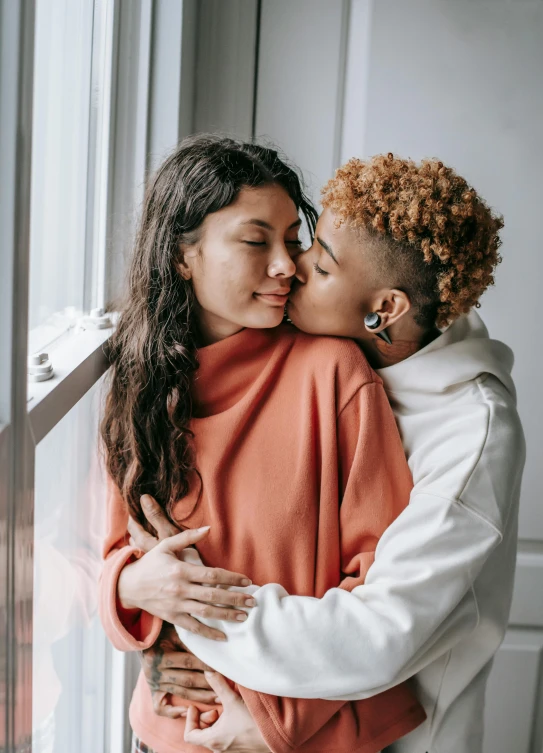 an adult woman kissing a younger woman's face while standing next to the window