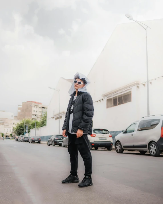 a man wearing all black standing on the street with a cloudy sky behind him