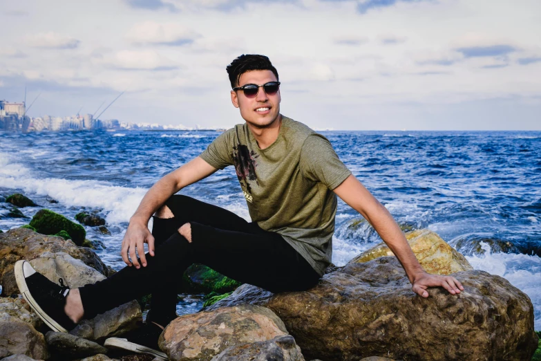 a man in sunglasses and a shirt sitting on a rock by the ocean