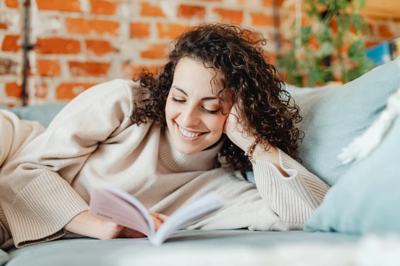 smiling woman in sweater lying on couch reading book