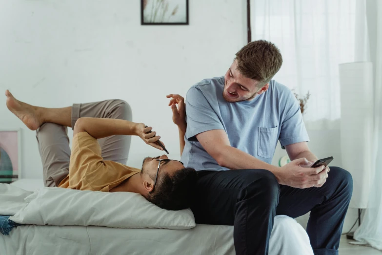 two men are sitting on a bed while using a mobile phone