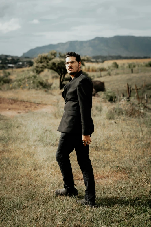 a young man dressed in a black suit stands on an open grassy field with mountains behind him