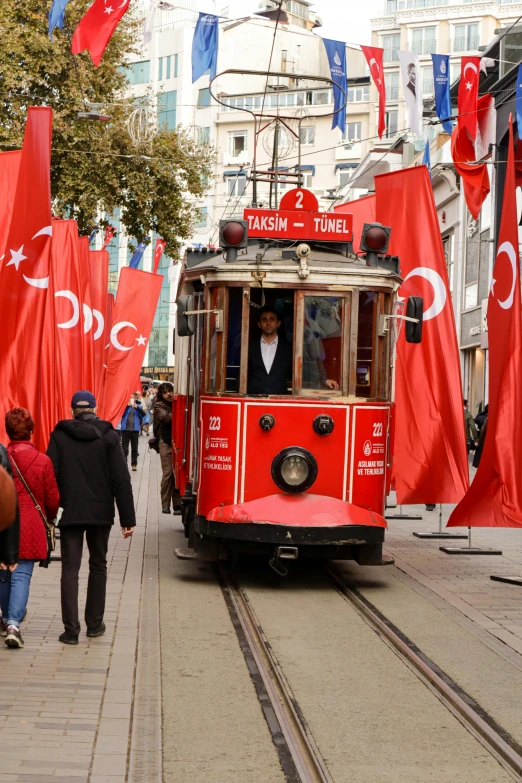 a red trolly car traveling down a road past many other people