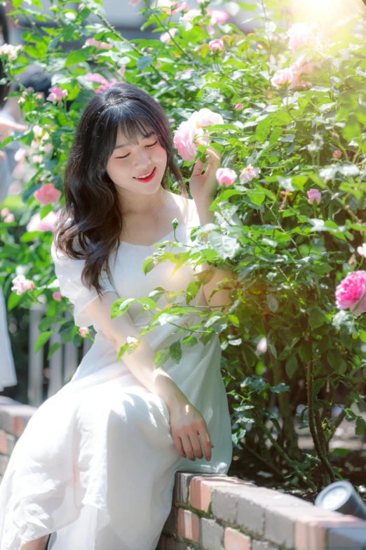 a young woman in white sitting on the side of a brick wall with flowers